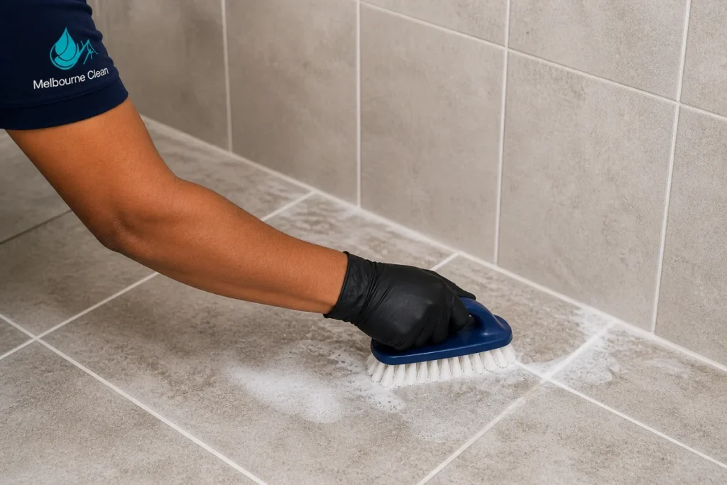 Home 26 A person wearing black gloves uses a scrub brush to clean light gray tiled surfaces.